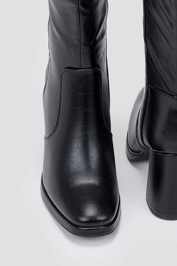 Close-up of black knee-high boot with a block heel and square toe, showing the shaft and heel on a light grey backdrop