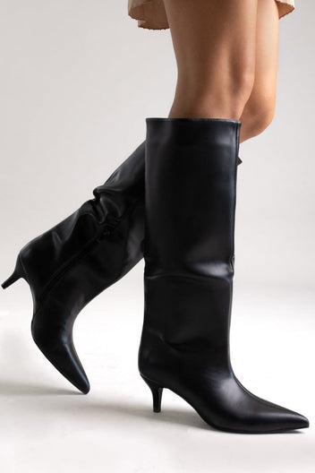 Model leg raised in black leather knee-high pointed toe boot with kitten heel, angled view on a light grey background