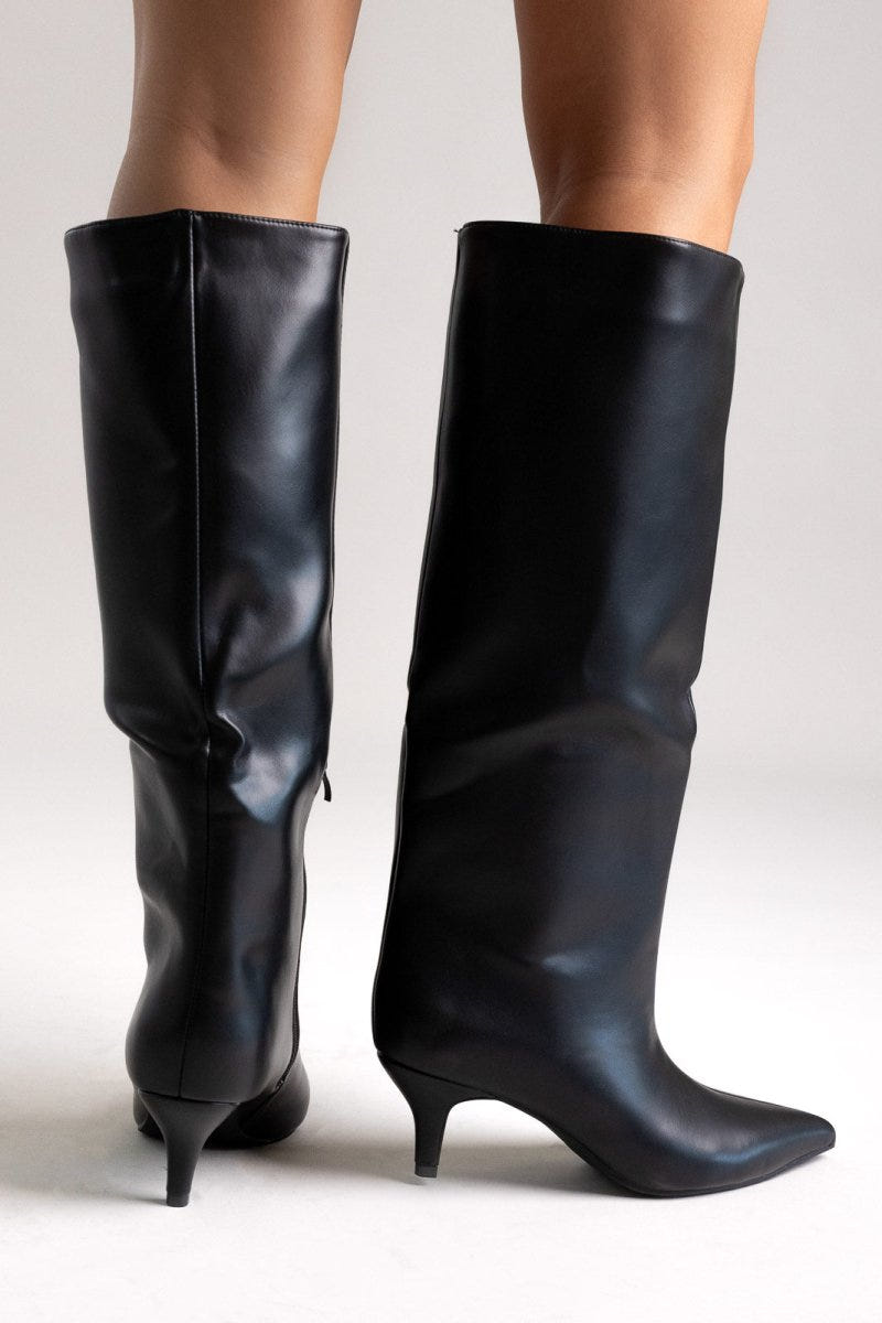 Pair of black leather knee-high pointed toe boots with slouchy shafts and kitten heels, angled view on light grey
