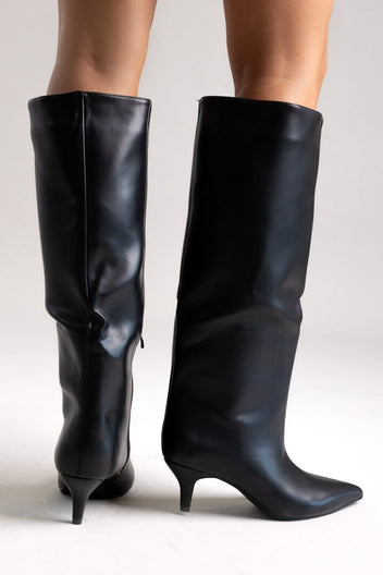 Pair of black leather knee-high pointed toe boots with slouchy shafts and kitten heels, angled view on light grey
