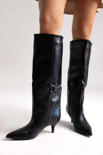 Pair of black leather knee-high boots with pointed toes, kitten heels and a metal buckle detail, angled view on light grey
