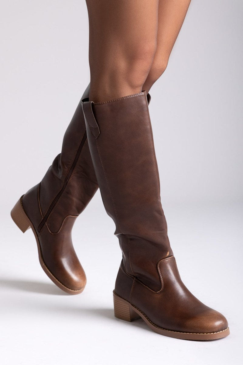 Model leg raised in brown leather knee-high boot with round toe and tan block heel, angled view on a light grey background