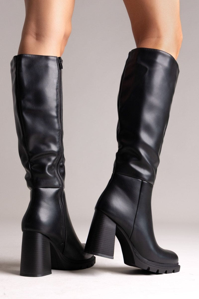 Model legs in black leather knee-high boots with block heels and lug soles, front view on a light grey background