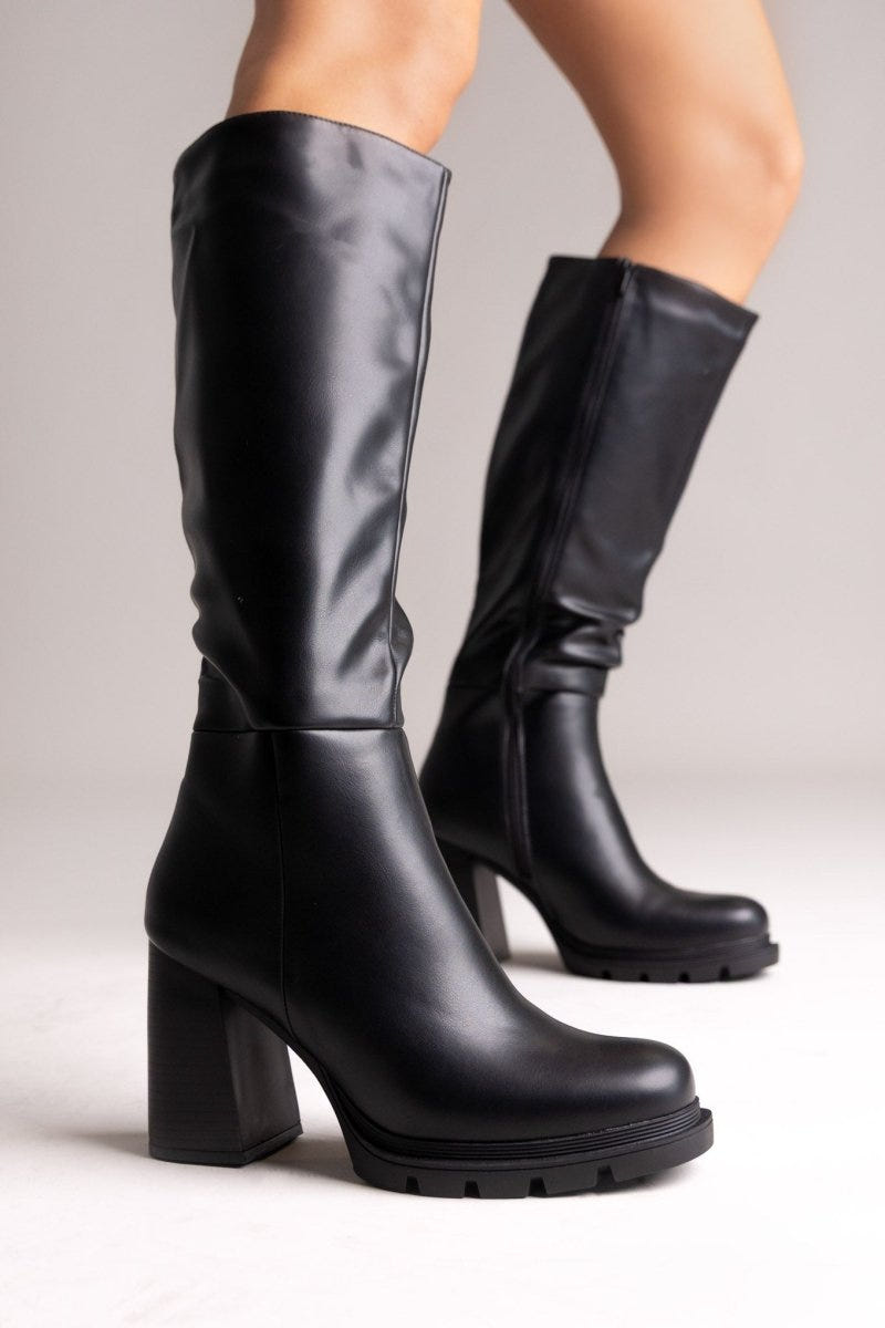 Model legs in black knee-high block heel boots with lug sole and side zip, side view on light grey background