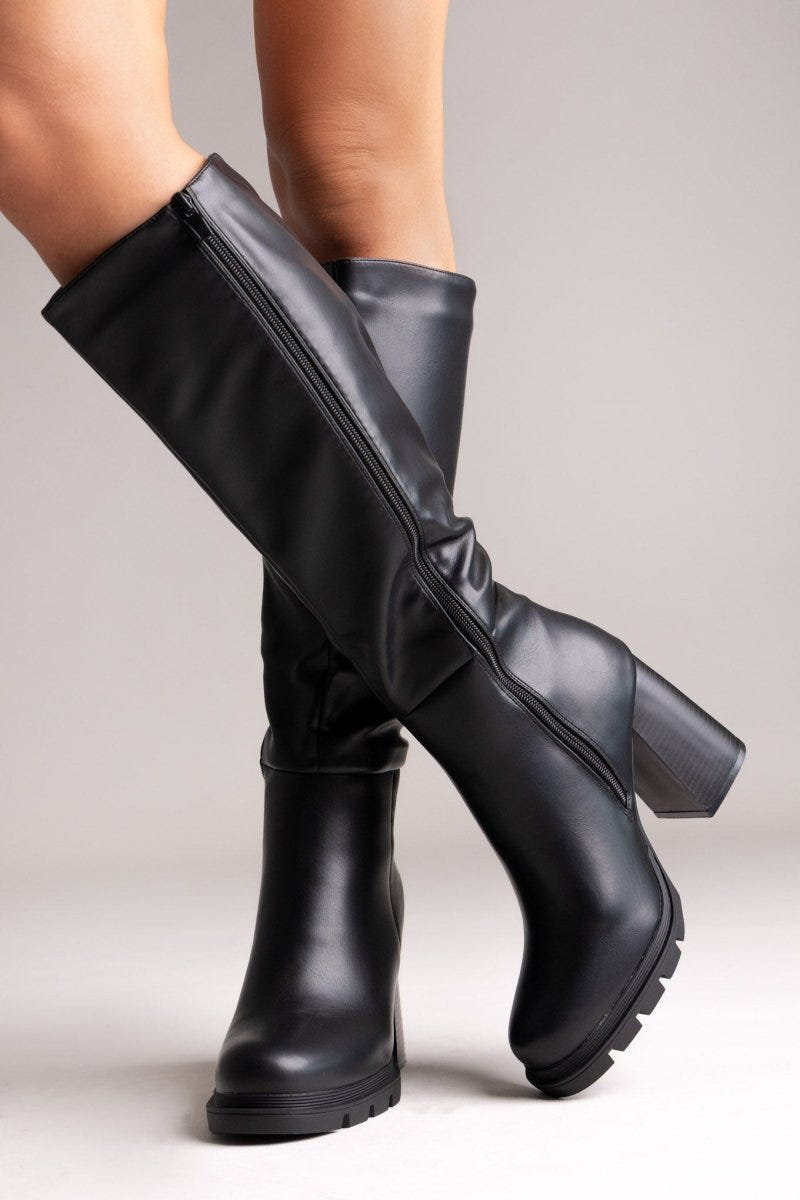 Model legs in black knee-high block heel boots with lug sole and buckle detail, angled view on light grey background