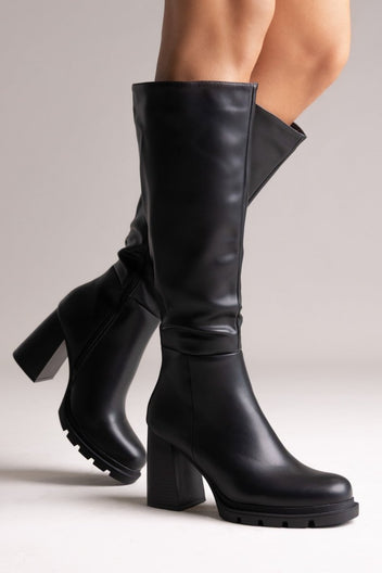 Model leg raised in black leather knee-high boot with lug sole and block heel, angled view on a light background