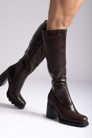 Model legs in dark brown knee-high platform block heel boots with lug sole, angled view on light grey background
