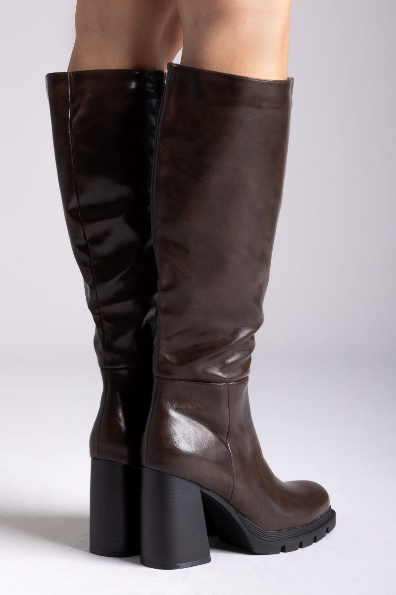 Pair of dark brown knee-high platform block heel boots with side zip and lug sole, front view on light grey background
