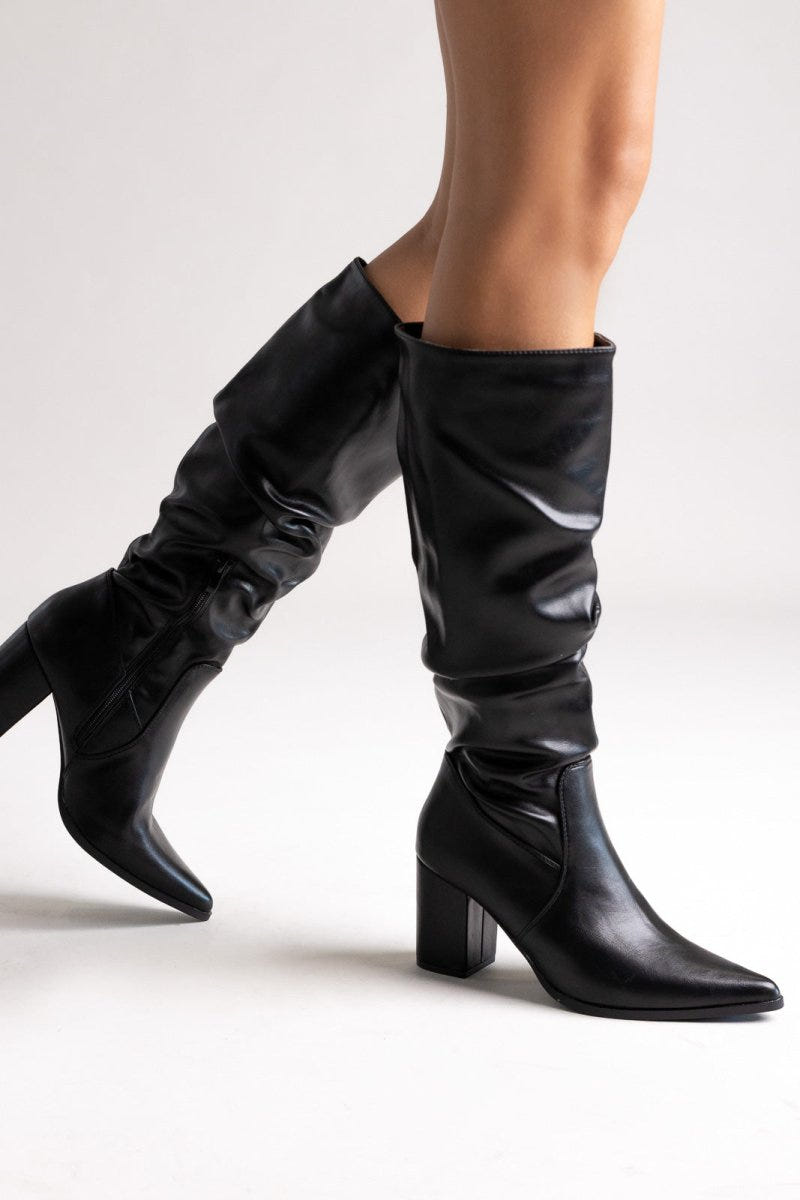 Model legs in black pointed toe ruched knee-high boots with block heel, angled view on light grey background