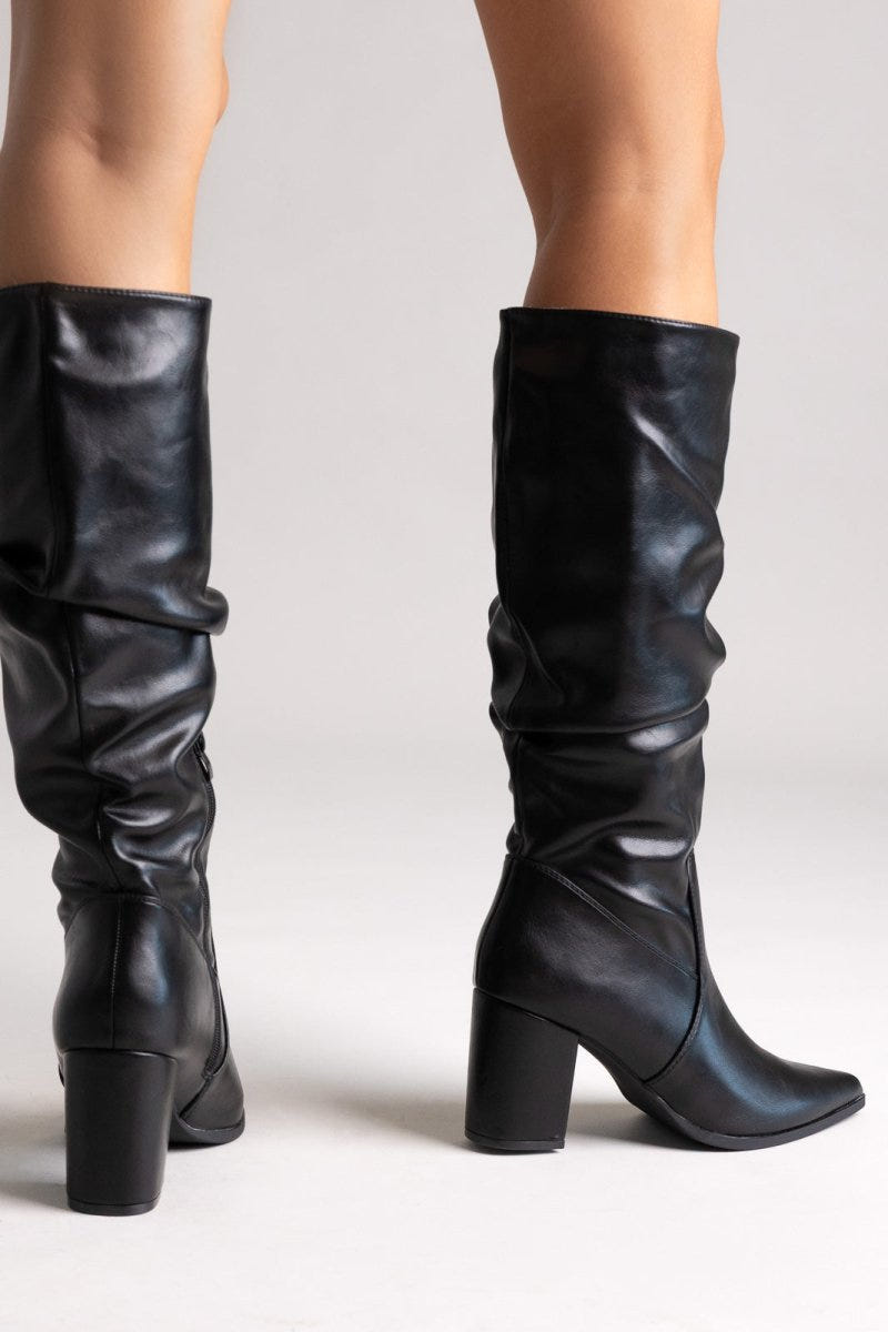 Model legs in black pointed toe ruched knee-high boots with block heel, front view on light grey background
