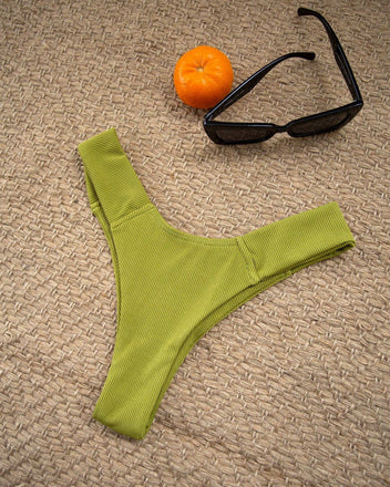 Lime green textured high-cut bikini bottom laid flat beside an orange and sunglasses on a woven beige mat