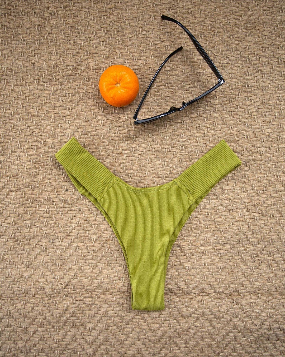 Lime green textured high-cut bikini bottom laid flat beside an orange and sunglasses on a woven beige mat