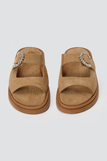 Pair of tan suede slide sandals with decorative silver buckle and chunky sole, overhead view on a light grey background