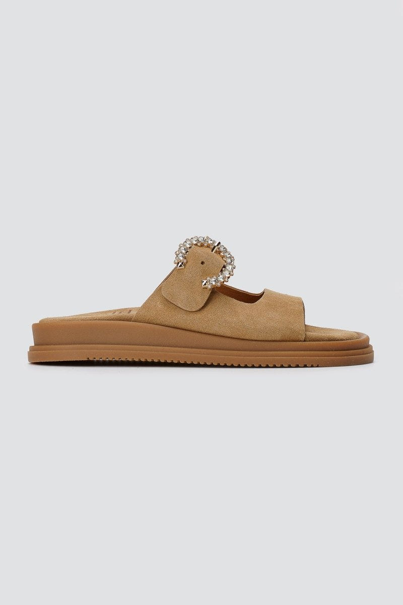 Tan suede slide sandal with decorative silver buckle and chunky sole, side view on a light grey background