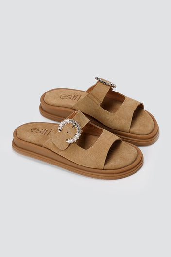 Pair of tan suede slide sandals with two decorative silver buckles and chunky sole, angled view on a light grey background