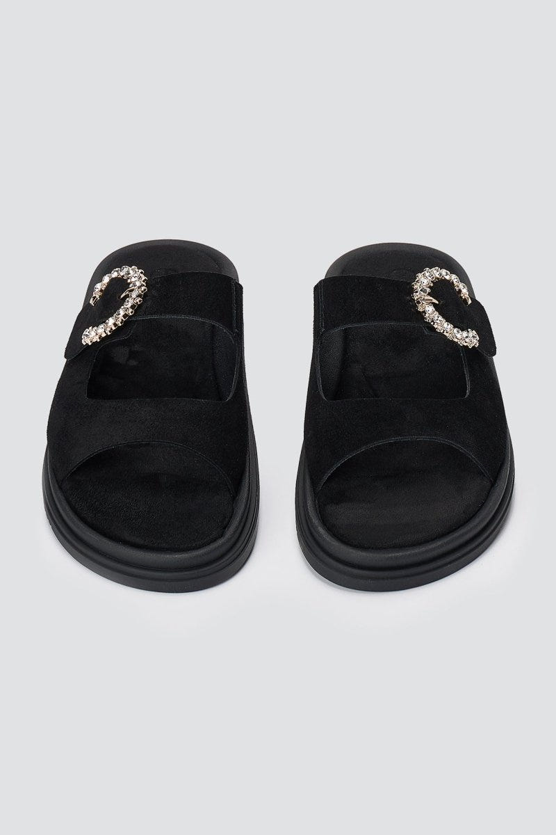 Pair of black suede slide sandals with decorative rhinestone buckle and chunky sole, overhead view on a light grey background