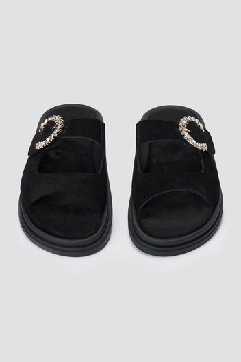Pair of black suede slide sandals with decorative rhinestone buckle and chunky sole, overhead view on a light grey background