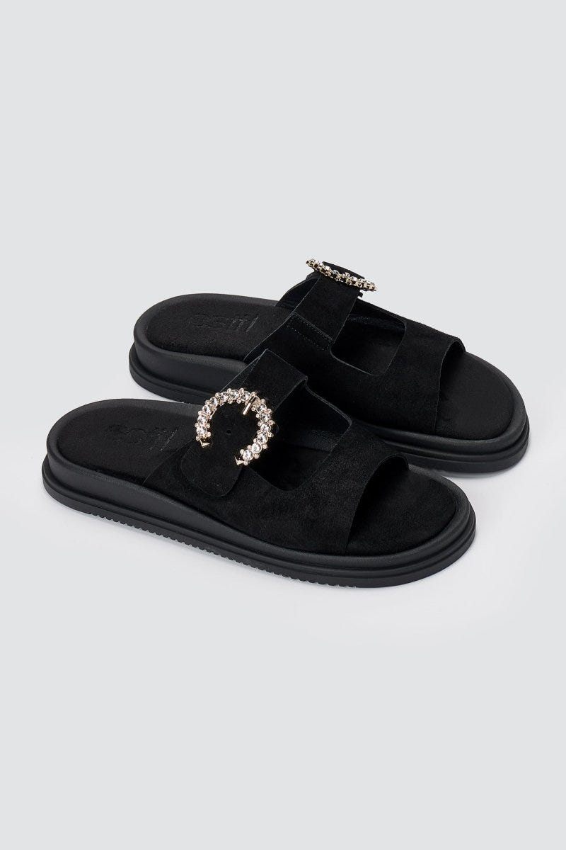 Pair of black suede slide sandals with decorative rhinestone buckle and chunky sole, angled view on a light grey background