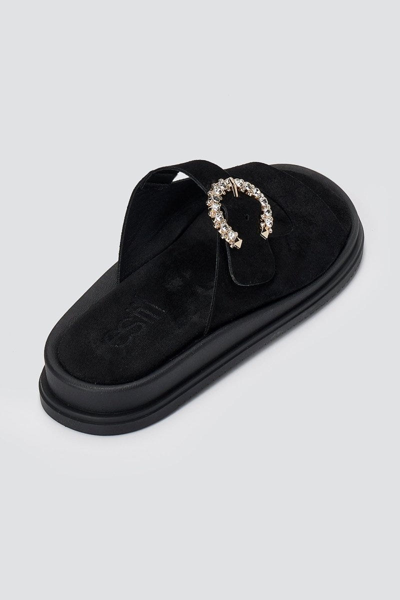 Black suede slide sandal with decorative rhinestone buckle and chunky sole, angled view on a light grey background