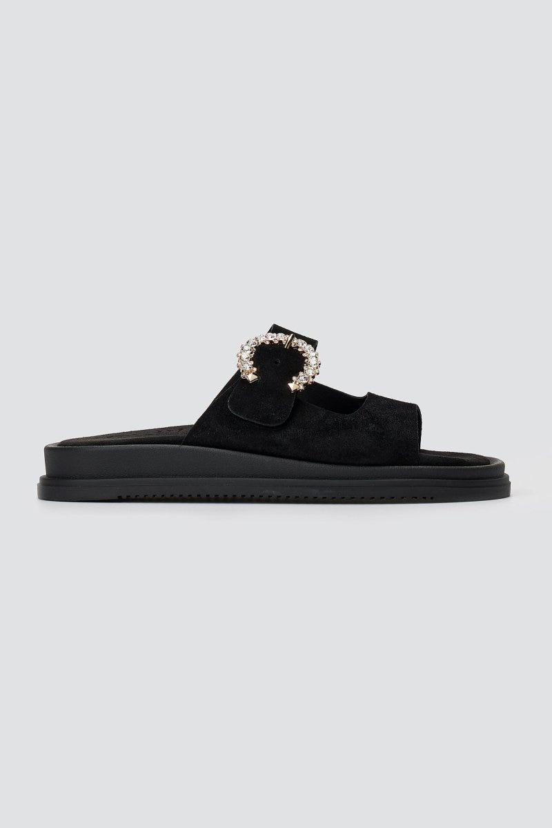 Black suede slide sandal with decorative buckle and chunky sole, side view on a light grey background