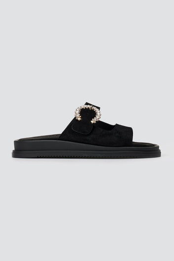 Black suede slide sandal with decorative buckle and chunky sole, side view on a light grey background