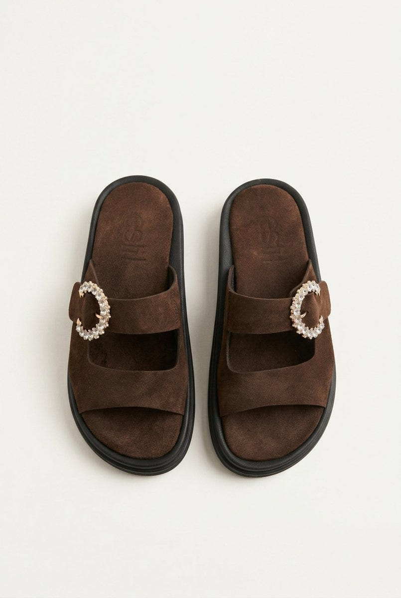 Pair of dark brown suede slide sandals with two straps and rhinestone buckle, overhead view on a light grey background