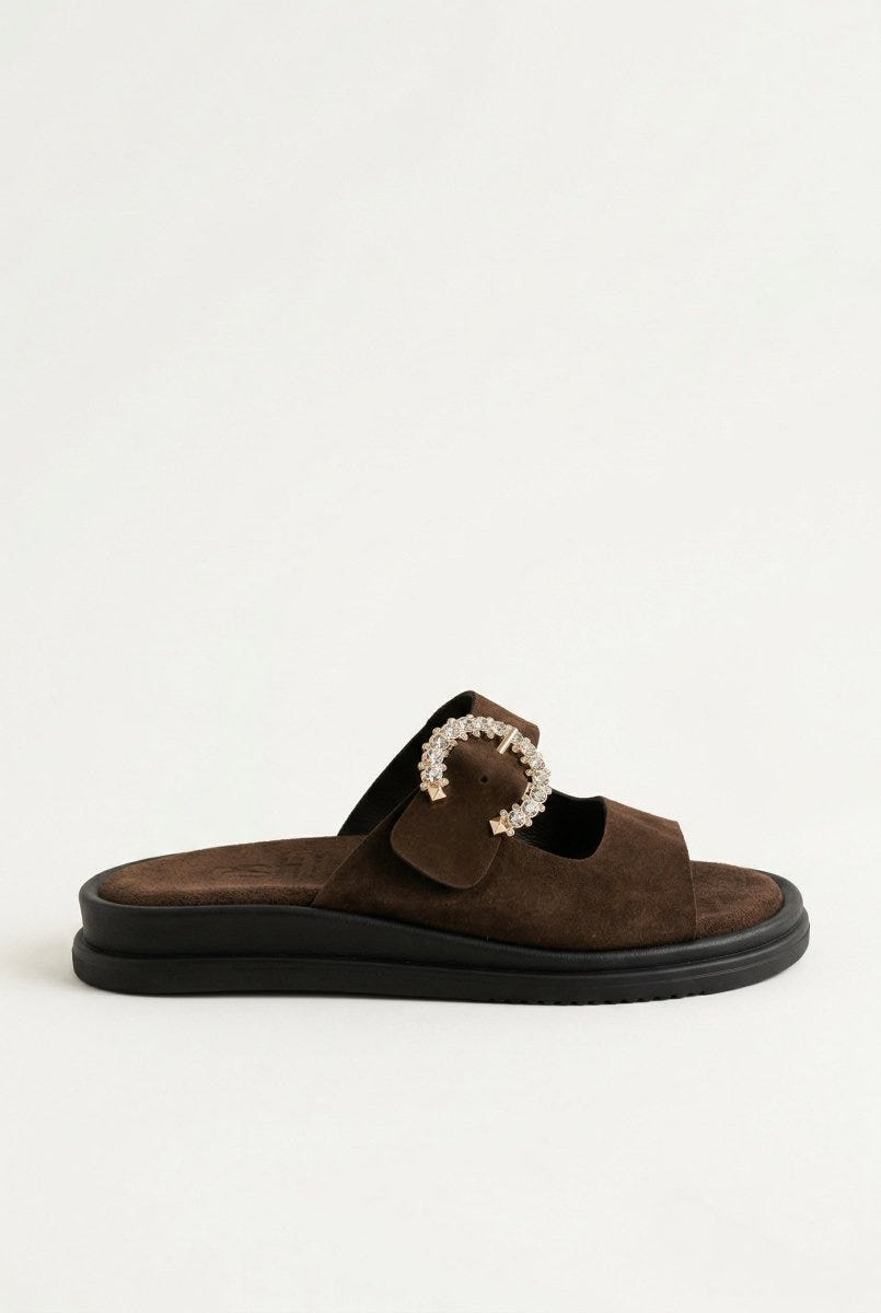 Dark brown suede slide sandal with decorative rhinestone buckle and dark chunky sole, angled view on a light grey background