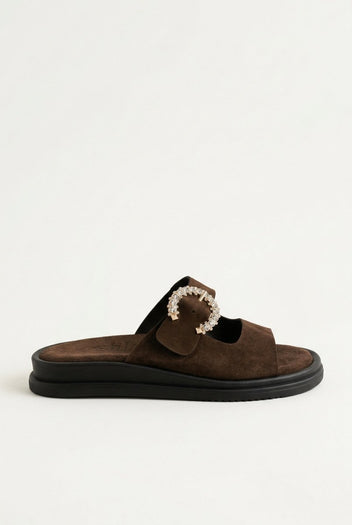 Dark brown suede slide sandal with decorative rhinestone buckle and dark chunky sole, angled view on a light grey background