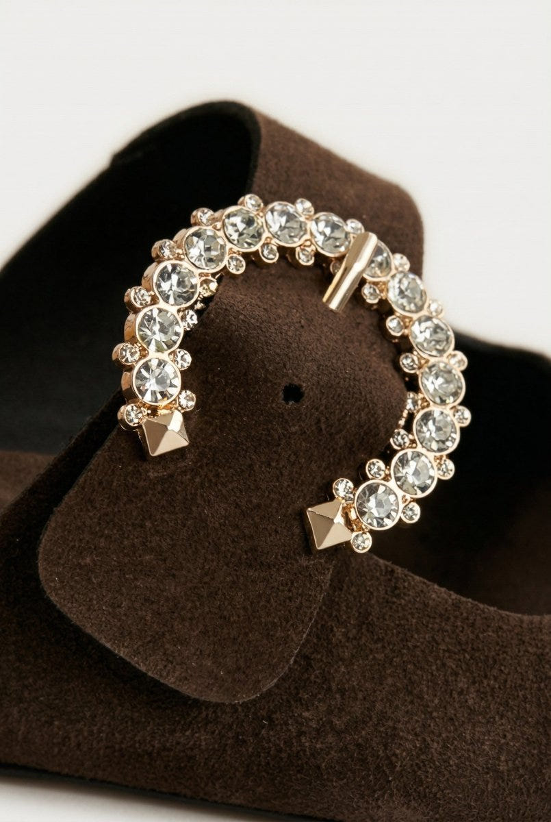Close-up of dark brown suede slide sandal with large rhinestone buckle on a light background