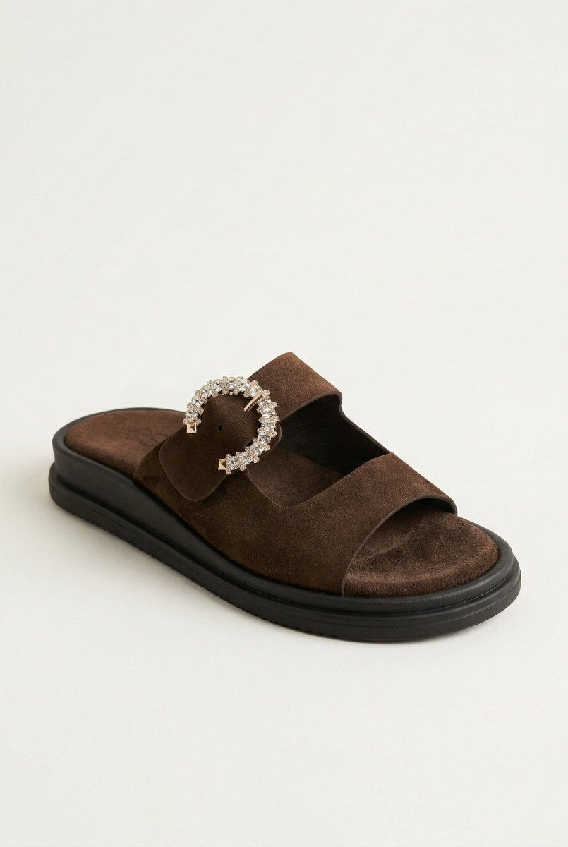 Dark brown suede slide sandal with two straps, rhinestone buckle and dark chunky sole, angled view on a light grey background