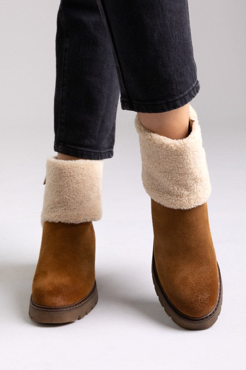 Model legs in tan suede shearling-cuff ankle boots with block heels and dark jeans, angled view on a light grey background