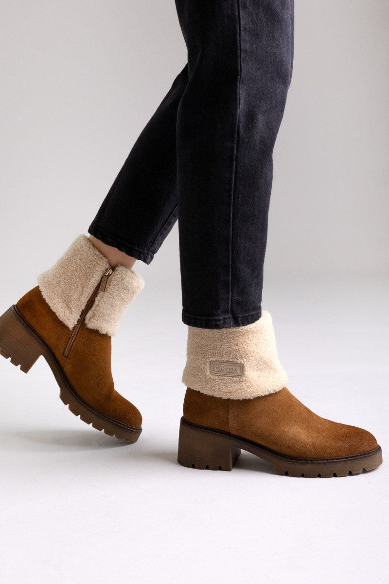 Model legs in tan suede shearling-cuff ankle boots with block heels and dark jeans, angled view on a light grey background