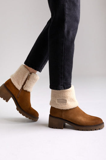 Model legs in tan suede shearling-cuff ankle boots with block heels and dark jeans, angled view on a light grey background