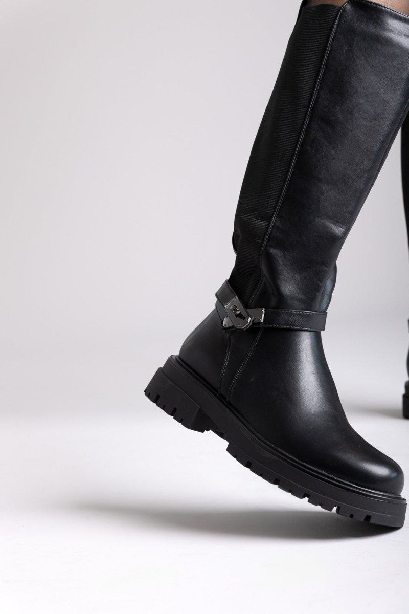 Model leg raised in black leather knee-high lug sole boot with buckle strap and rounded toe, angled view on a light background