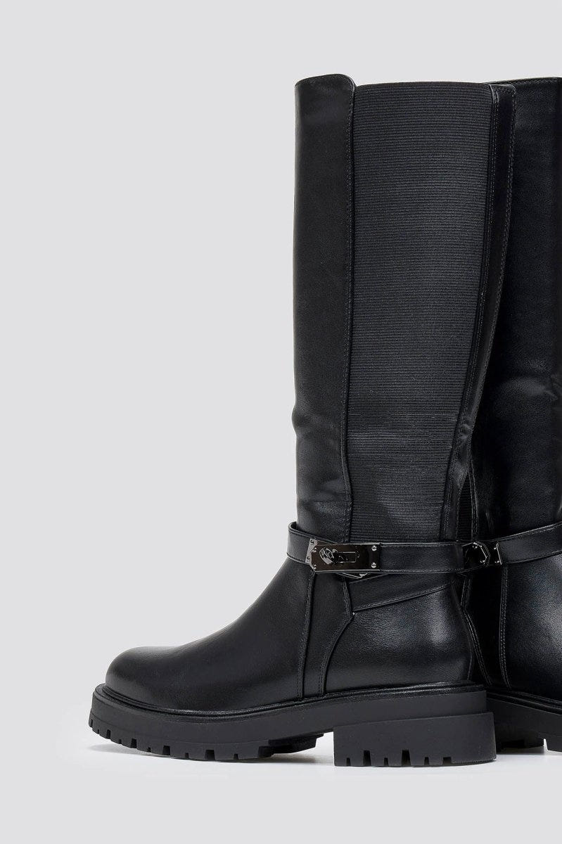 Pair of black leather knee-high lug sole boots with buckle strap at the ankle, angled view on a light grey background