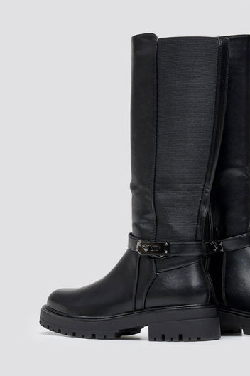 Pair of black leather knee-high lug sole boots with buckle strap at the ankle, angled view on a light grey background