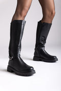Model legs in black leather knee-high lug sole boots with buckle strap and side zip, front view on a light background