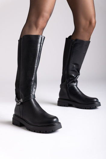 Model legs in black leather knee-high lug sole boots with buckle strap and side zip, front view on a light background