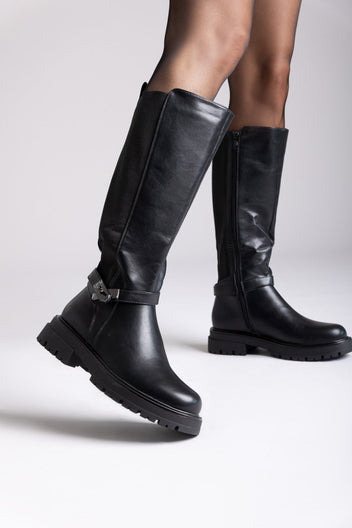 Model legs in black leather knee-high lug sole boots with buckle strap, angled view on a light background