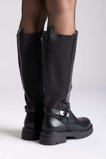 Model legs in black leather knee-high lug sole boots with buckle strap and side zip, angled view on a light background