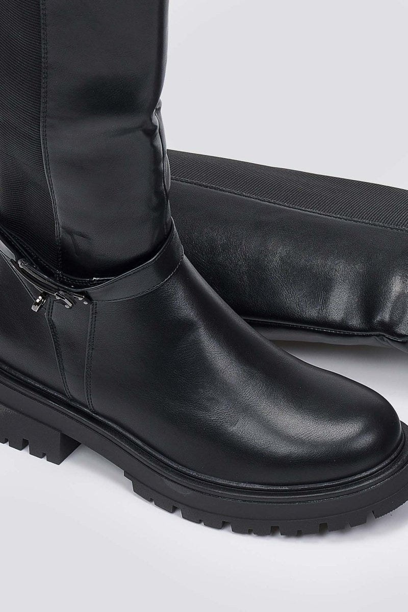 Close-up of black leather lug sole boot with buckle strap and rounded toe on a light grey background