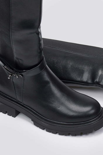 Close-up of black leather lug sole boot with buckle strap and rounded toe on a light grey background