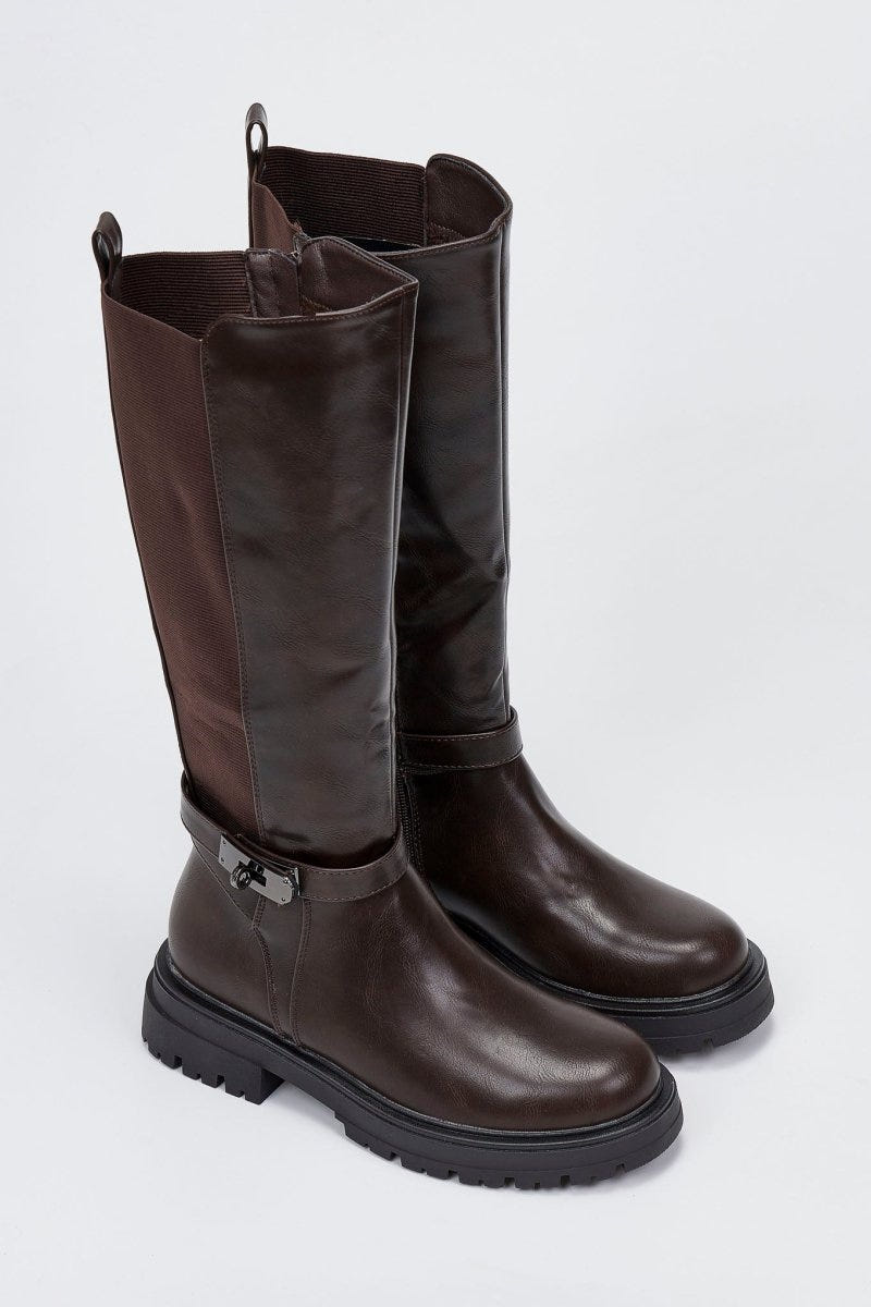 Pair of dark brown and black knee-high lug sole boots with buckle strap, angled view on a light grey background