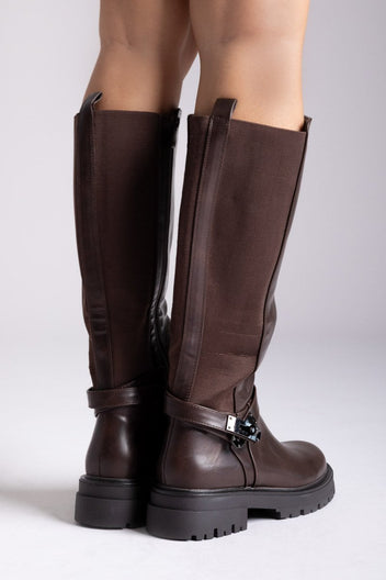 Model legs in dark brown knee-high lug sole boots with buckle strap, front view on a light grey background