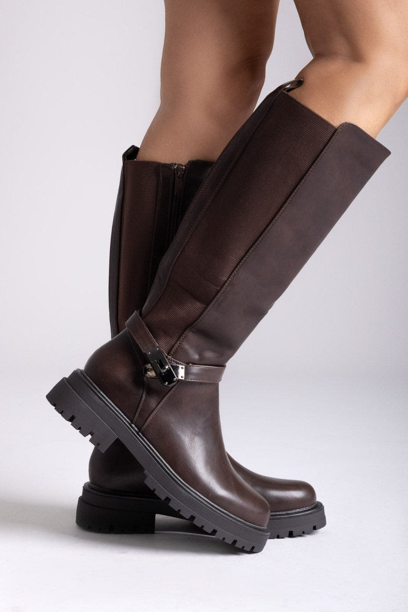 Model legs in dark brown knee-high lug sole boots with buckle strap and stretch panel, angled view on a light background