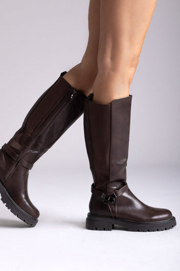 Model leg raised in dark brown knee-high lug sole boot with buckle strap and stretch panel, angled view on a light background