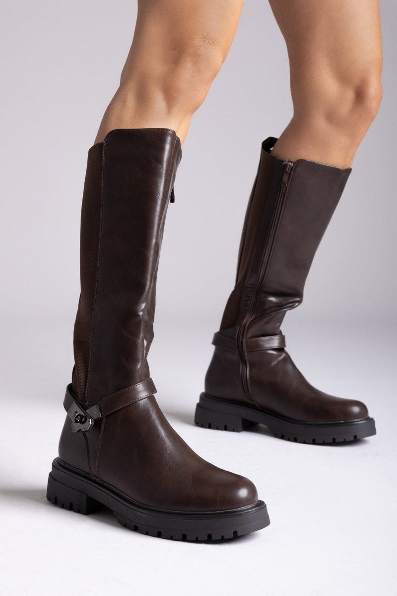 Model legs in dark brown knee-high lug sole boots with buckle strap and side zip, angled view on a light grey background