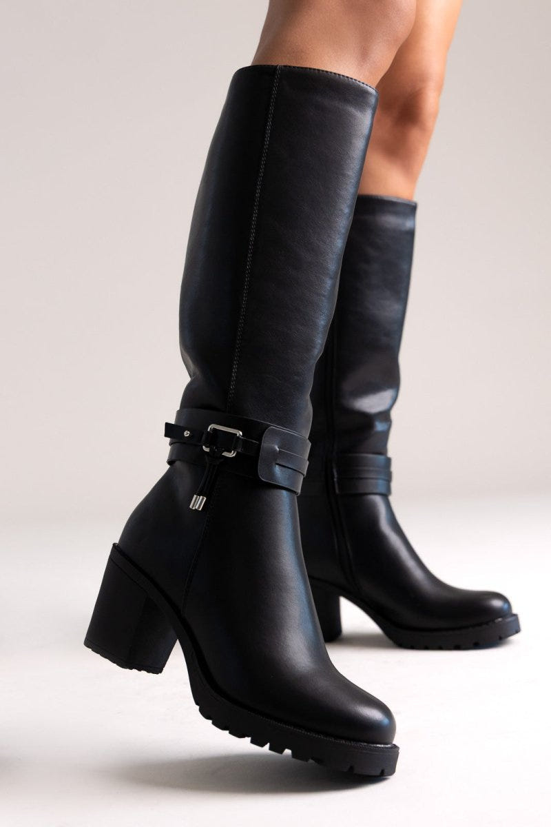 Model legs in black leather knee-high block heel boots with buckle strap and side zip, angled view on a light background