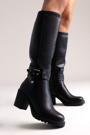 Model legs in black leather knee-high block heel boots with buckle strap and side zip, angled view on a light background