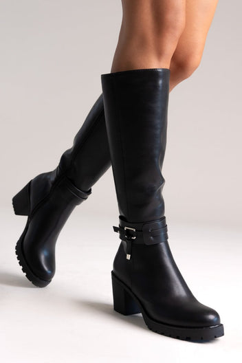 Model leg raised in black leather knee-high block heel boot with buckle strap and lug sole, angled view on a light background